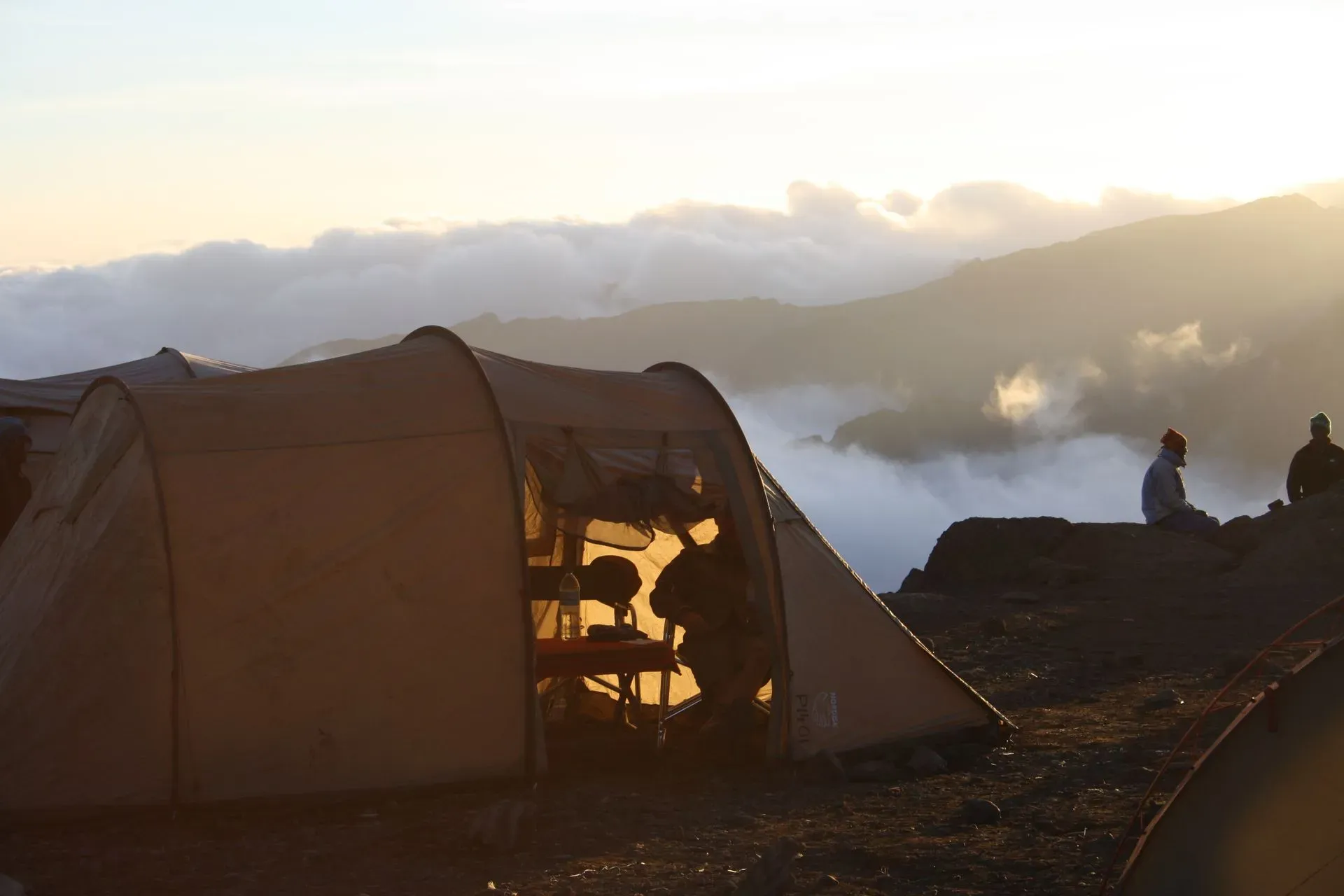 People sitting in tents on Mount Kilimanjaro, surrounded by rocky terrain and a clear blue sky.